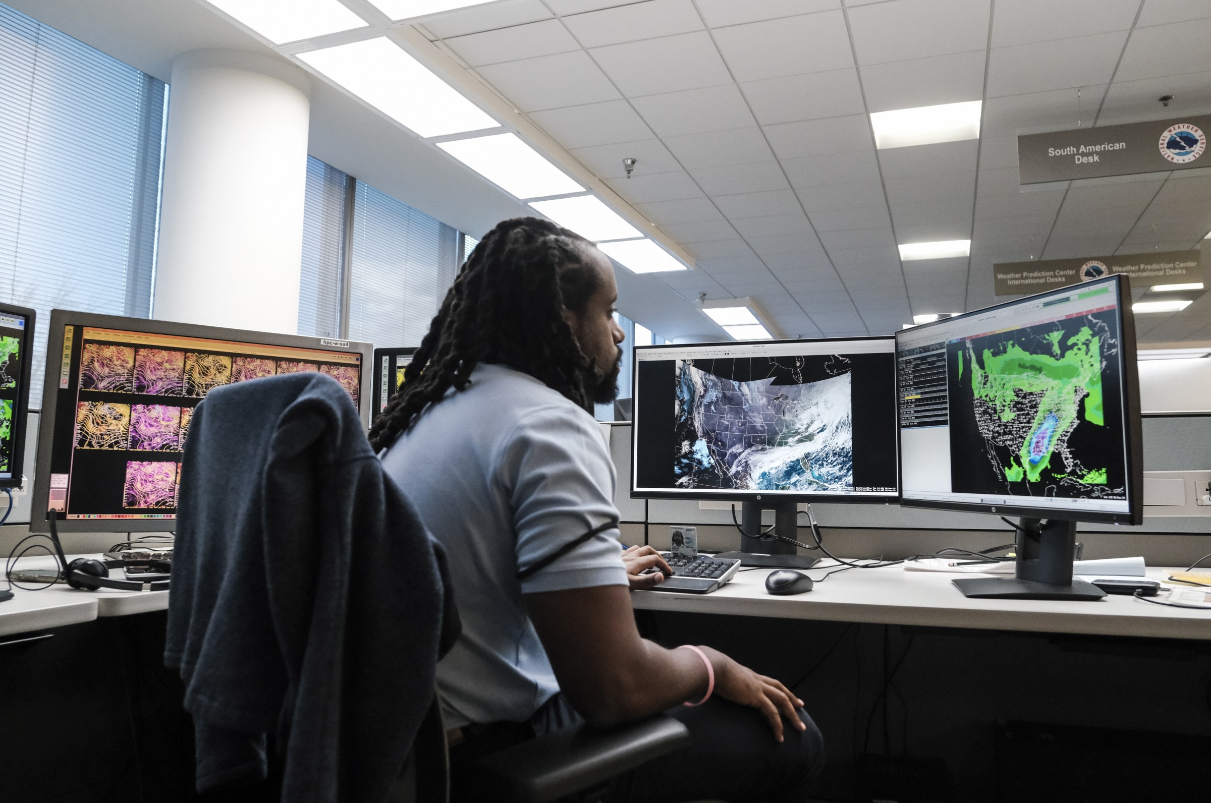 A man at a desk looks at several computer screens with maps of the US and weather blobs over the map.